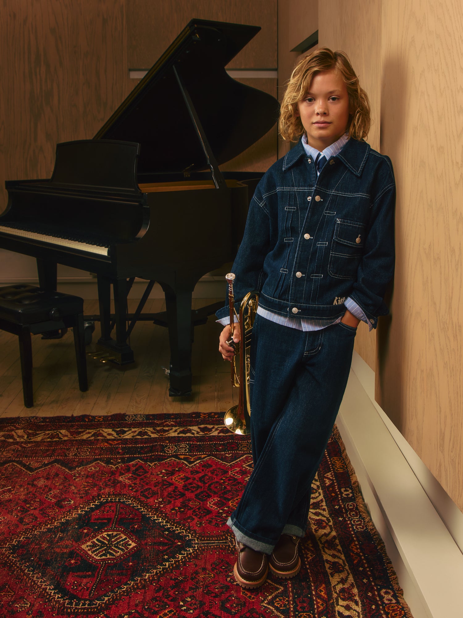 Blond boy in dark indigo Kith denim jacket and wide-leg jeans holding a brass trumpet by a grand piano on a red rug.