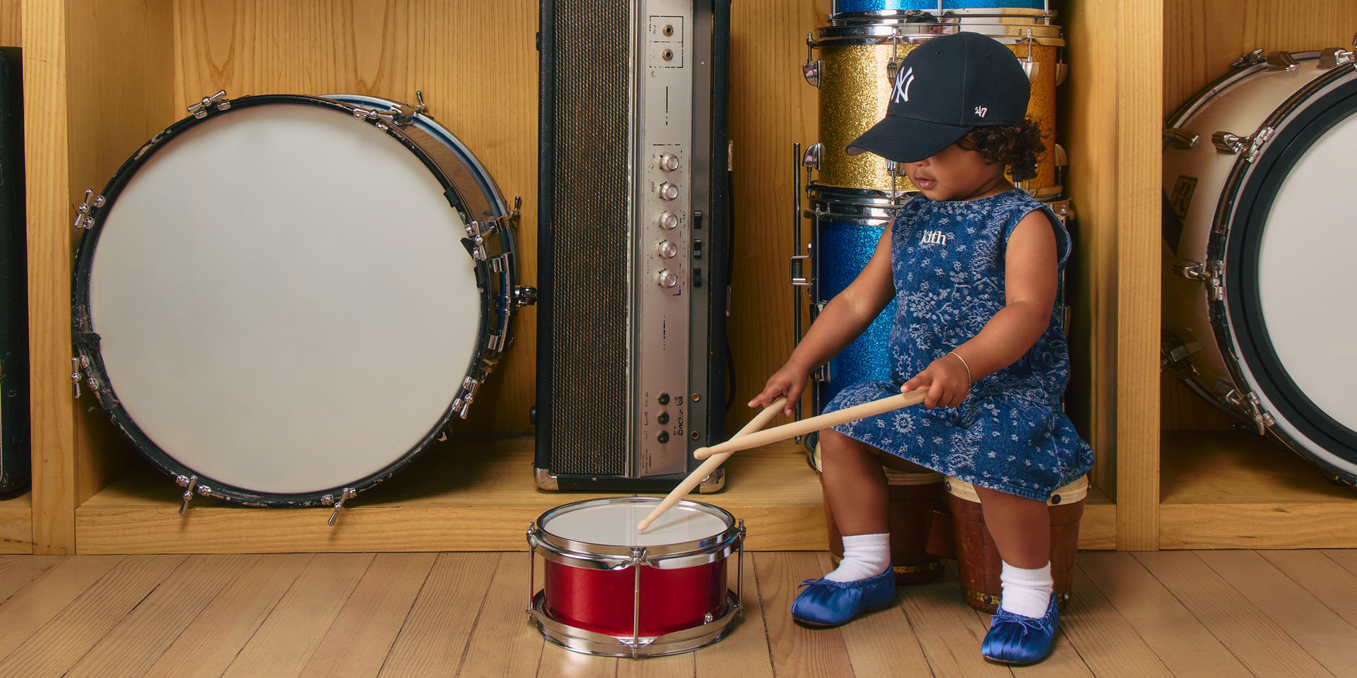 Toddler in Yankees cap and blue bandana-print Kith dress drumming a red toy snare with sticks, glitter drums on wood shelves.
