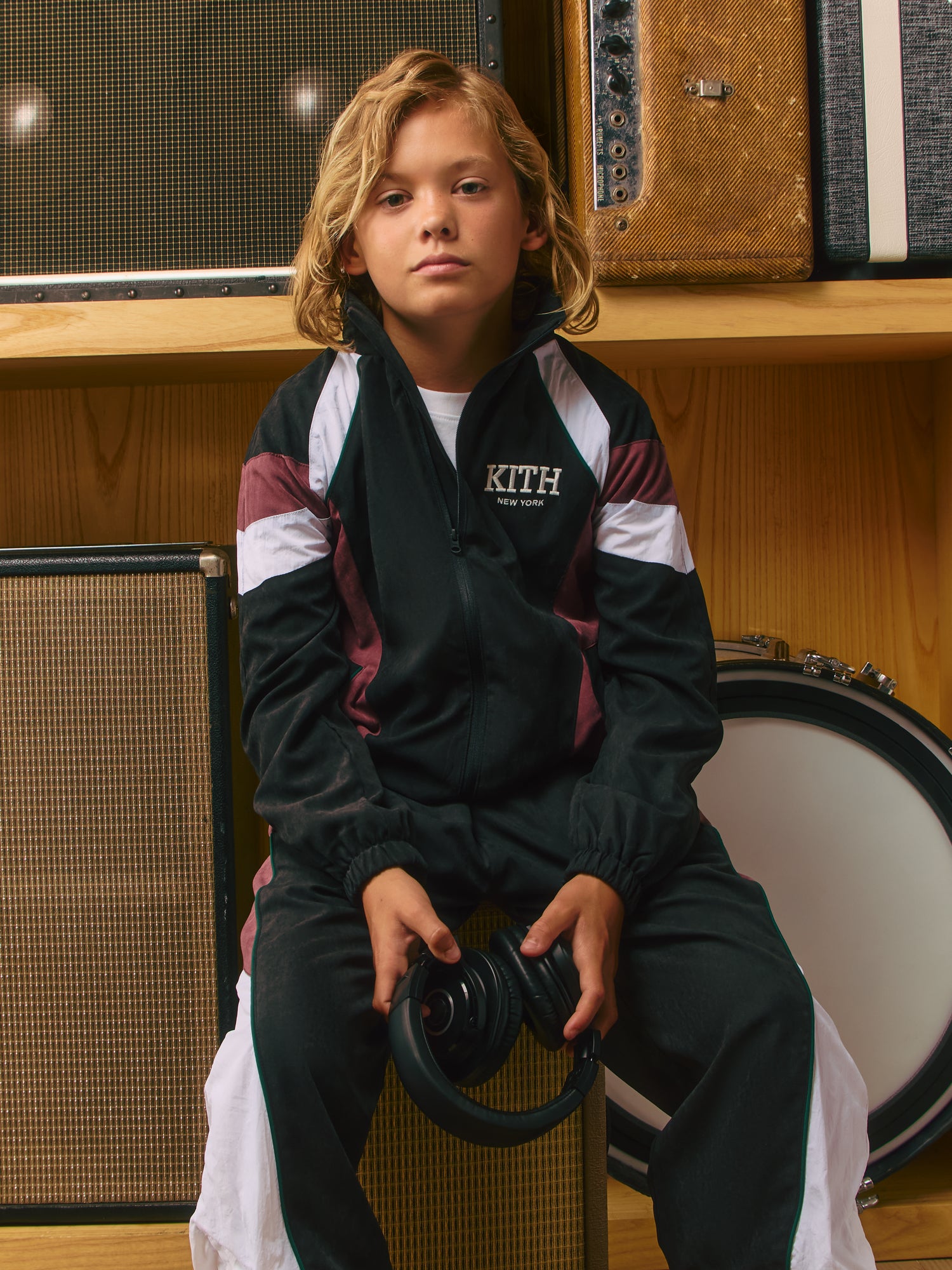 Blond boy in black KITH New York velour track jacket with maroon stripes holding headphones, amps and a drum behind.