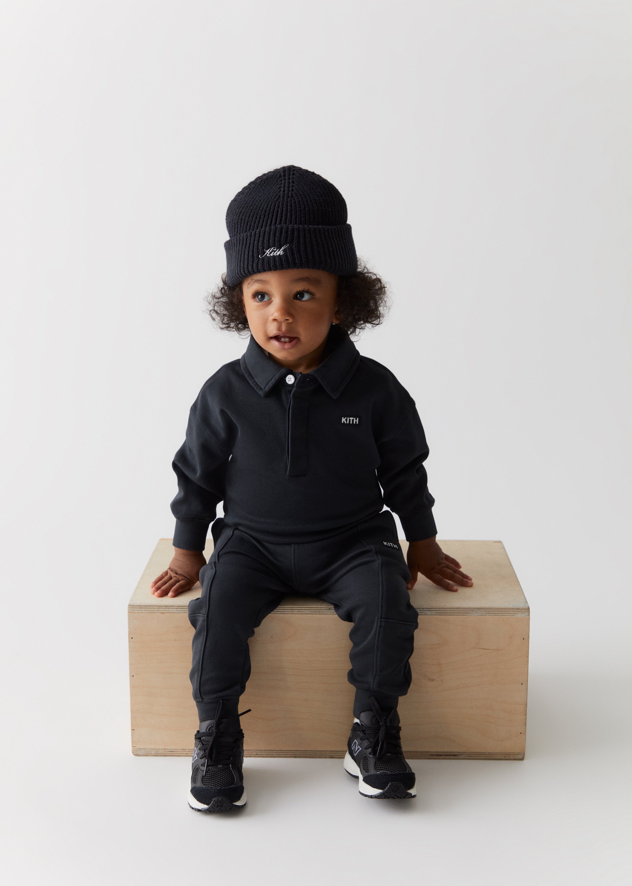 A toddler wearing a Kith script embroidered knit beanie, black long sleeve polo, and matching black pants from Kith Kids Fall Classics 2023.