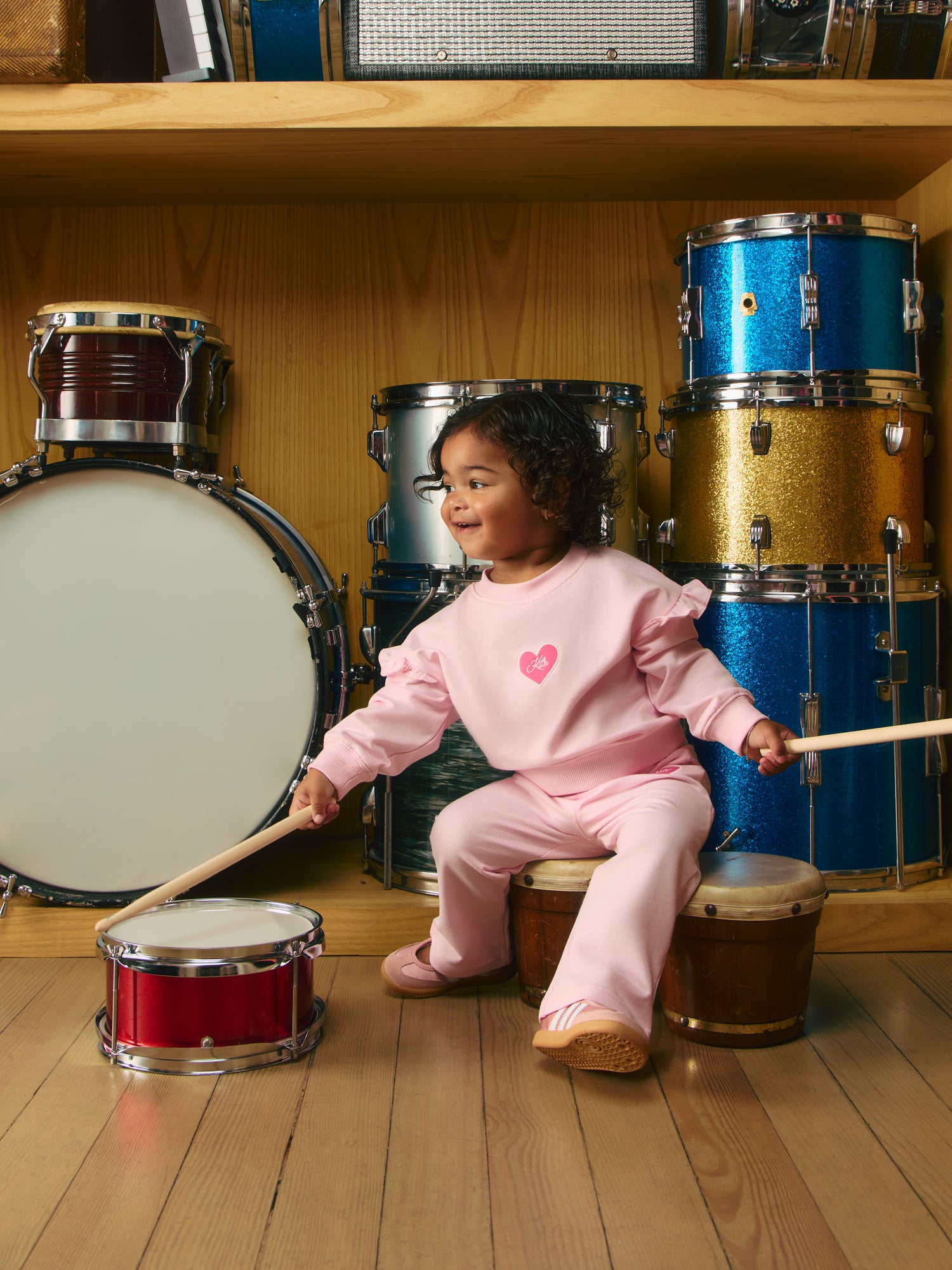 Curly-haired toddler in pink Kith ruffle sweatsuit with heart logo playing a red toy snare drum, glittered drum set behind.