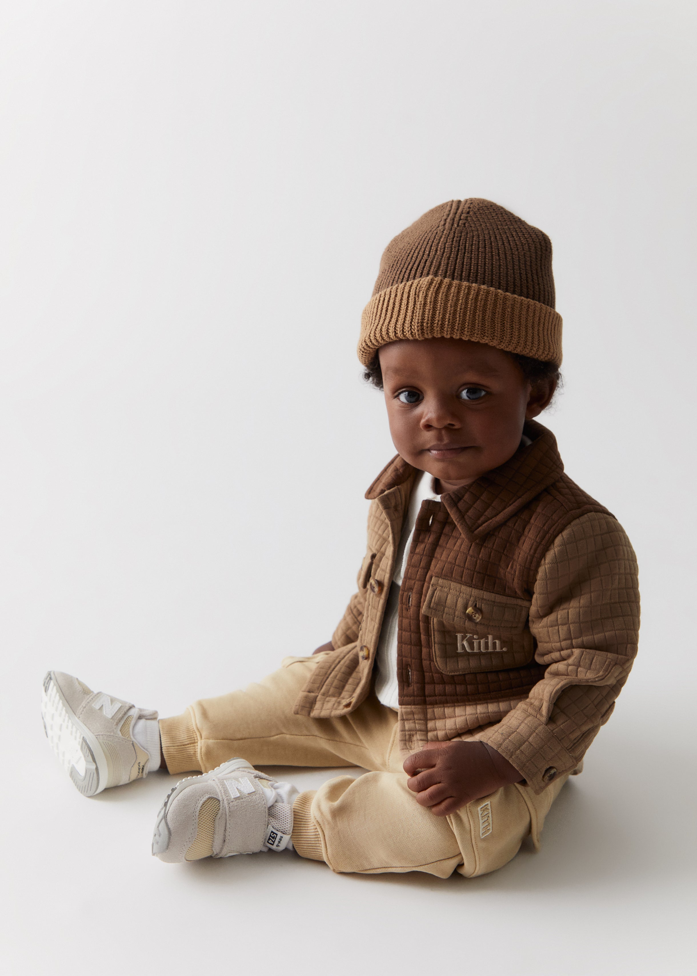 A baby sitting while wearing a knit brown beanie, a quilted brown jacket with Kith embroidery, and tan cargo pants, from the Kith Kids Fall Classics 2023 collection.