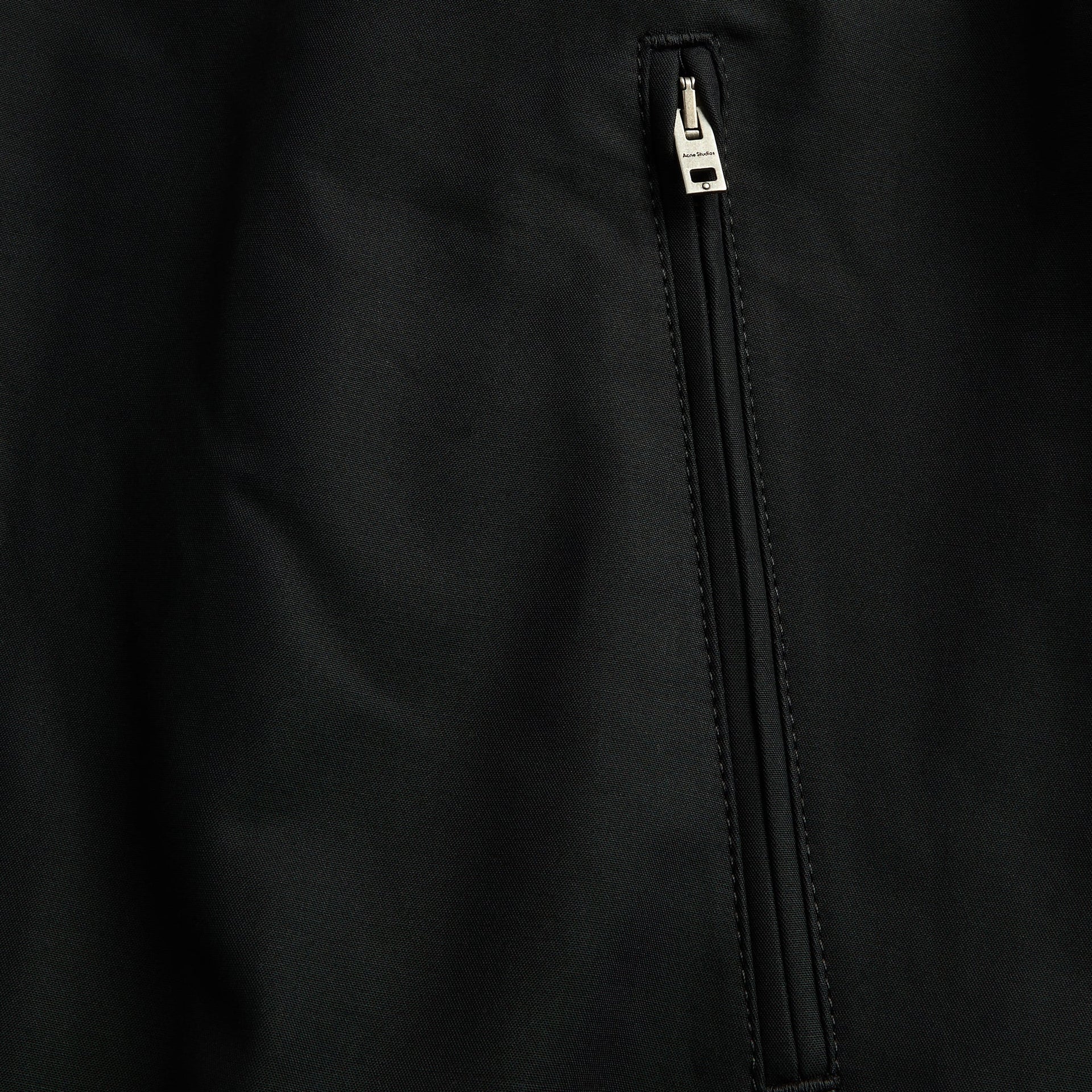 Acne Studios black bomber jacket detail, close-up of zip pocket pull with embossed branding