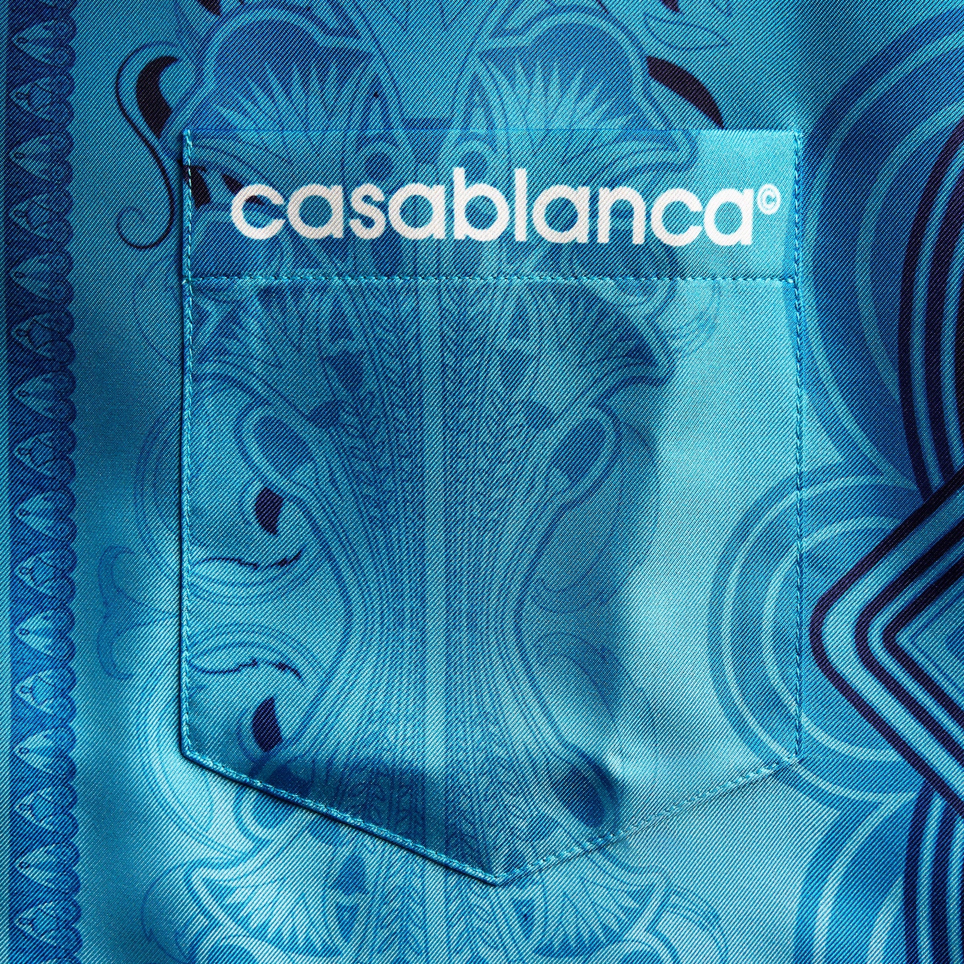 Close-up of blue Gradient Egyptian Rug silk: chest pocket with white casablanca logo over ornate scrollwork print