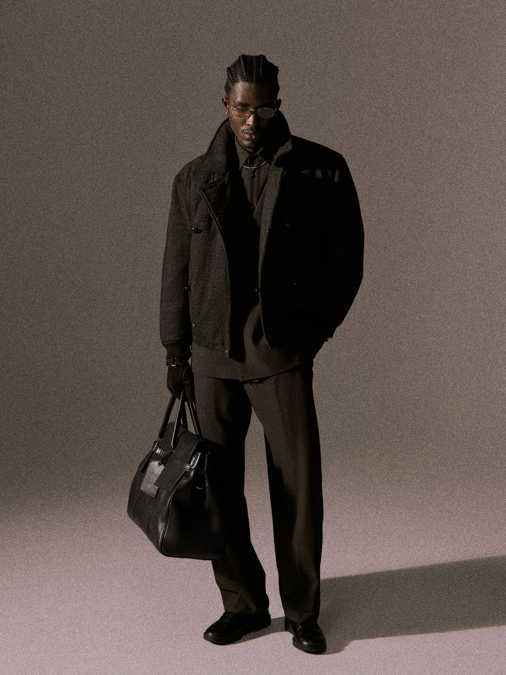 Kith Winter lookbook: model in sunglasses, black wool bomber jacket and trousers, holding black leather duffel, full-length