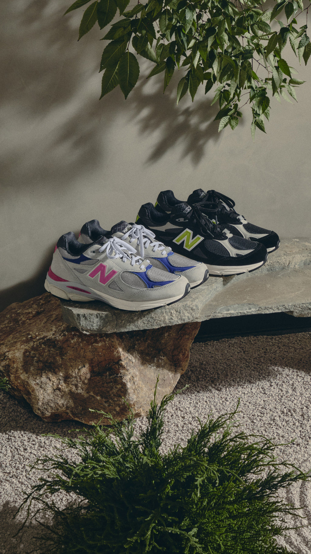 Pair of grey New Balance sneakers with pink N logo and a black pair with lime N logo on a stone slab under leaves, vertical view