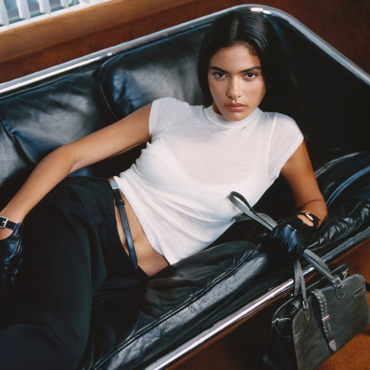 Model reclining on a black leather bench seat wearing a white t-shirt and black trousers, holding a black leather bag, close crop