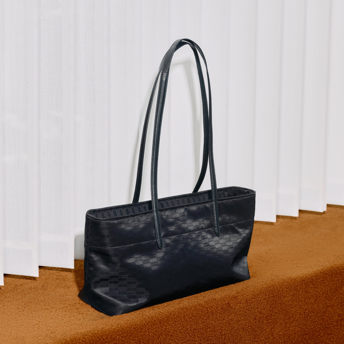 Black nylon tote bag with a woven Kith monogram pattern, leaning against white window blinds