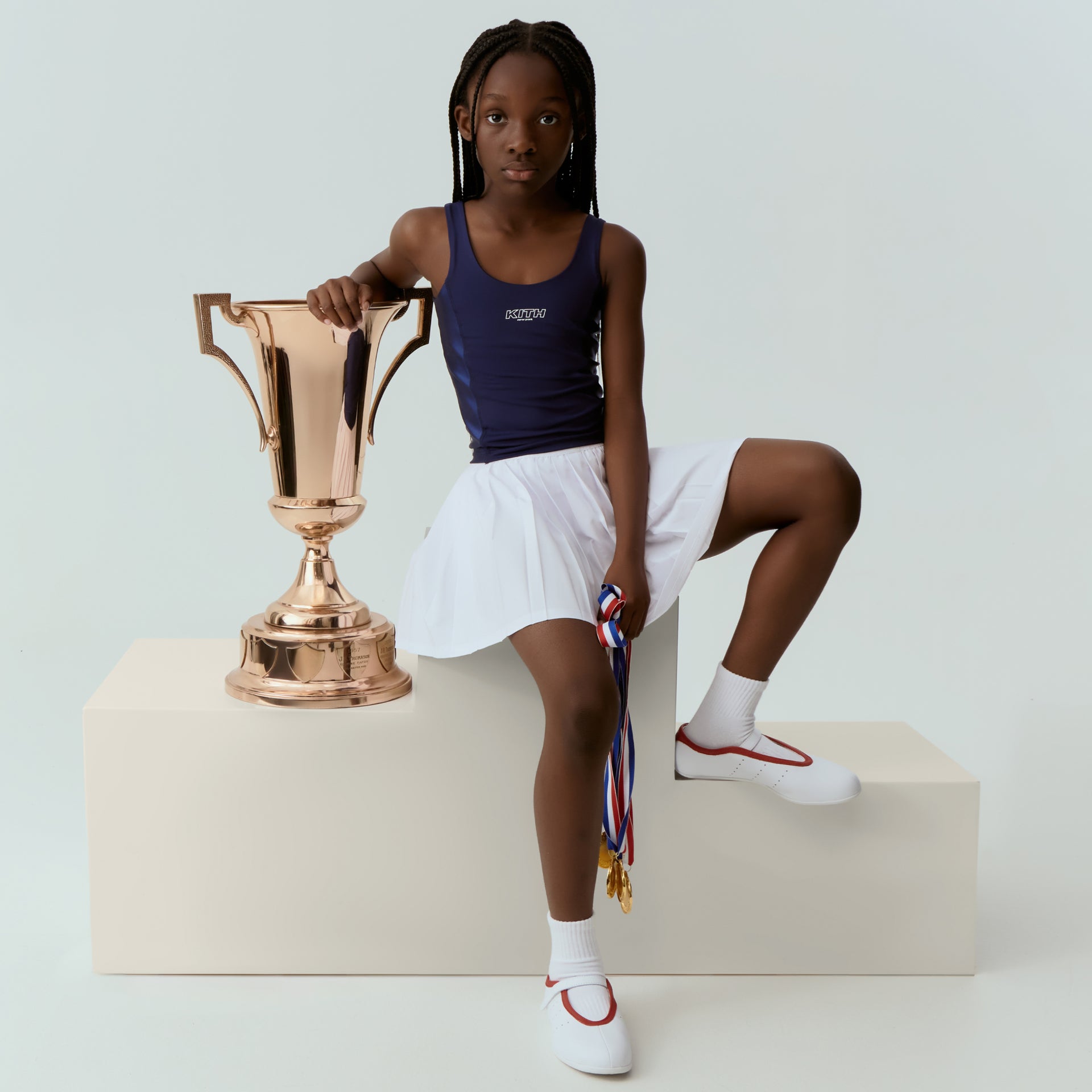 Girl in a navy Kith tank and white tennis skirt leans on a large rose-gold trophy holding medals, seated on a cream pedestal