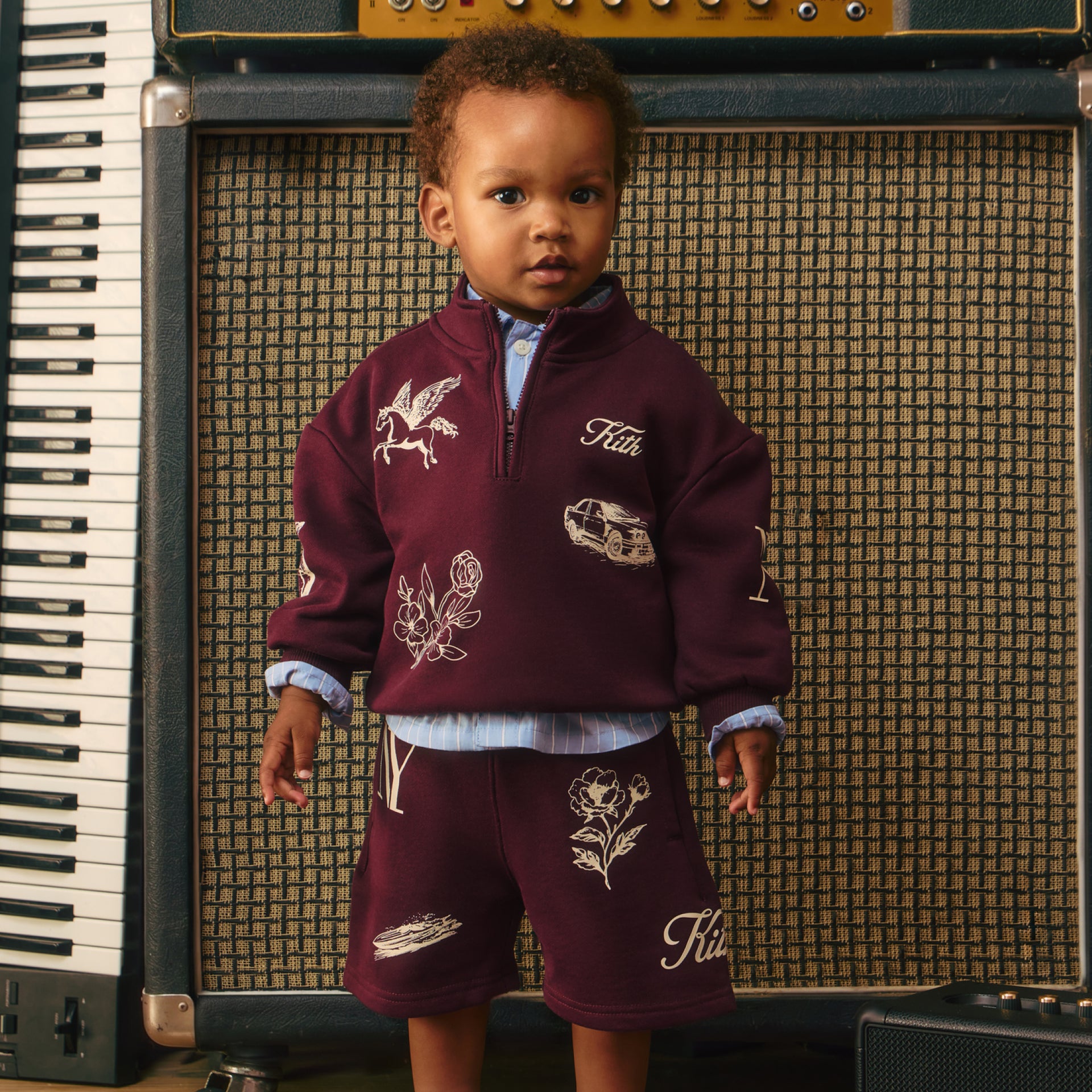 Toddler in a burgundy Kith quarter-zip and shorts with cream embroidered motifs and blue striped shirt, before a vintage guitar amp