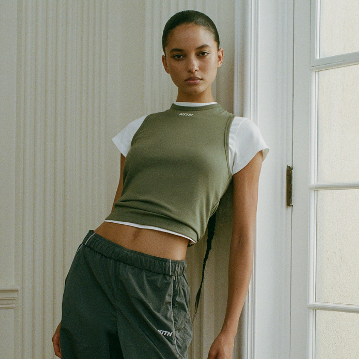 Model wearing an olive green sleeveless Kith top layered over a white t-shirt with dark cargo pants
