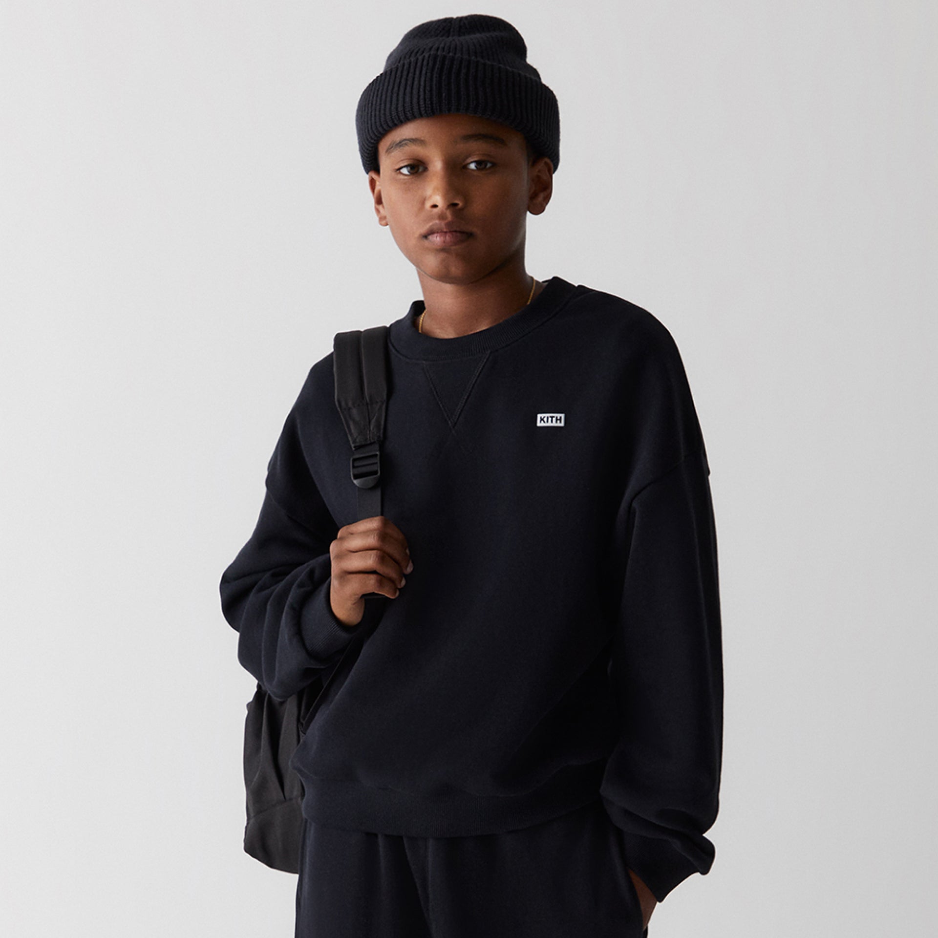 Blond kid in a white Kith K cap and gray rose polo sweatshirt sits against a vintage amp, cream New Balance sneakers, checked backpack