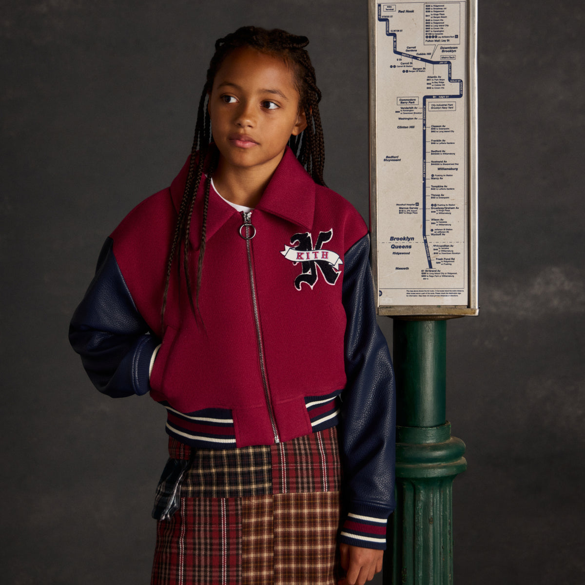 Girl in a burgundy and navy Kith varsity jacket with K logo patch and plaid patchwork skirt, standing next to a Brooklyn bus route sign