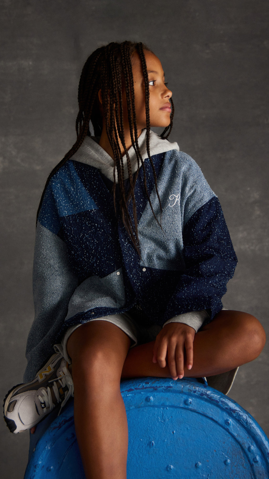 Girl in a speckled blue colorblock Kith cardigan over a grey hoodie, seated on a blue barrel wearing New Balance sneakers