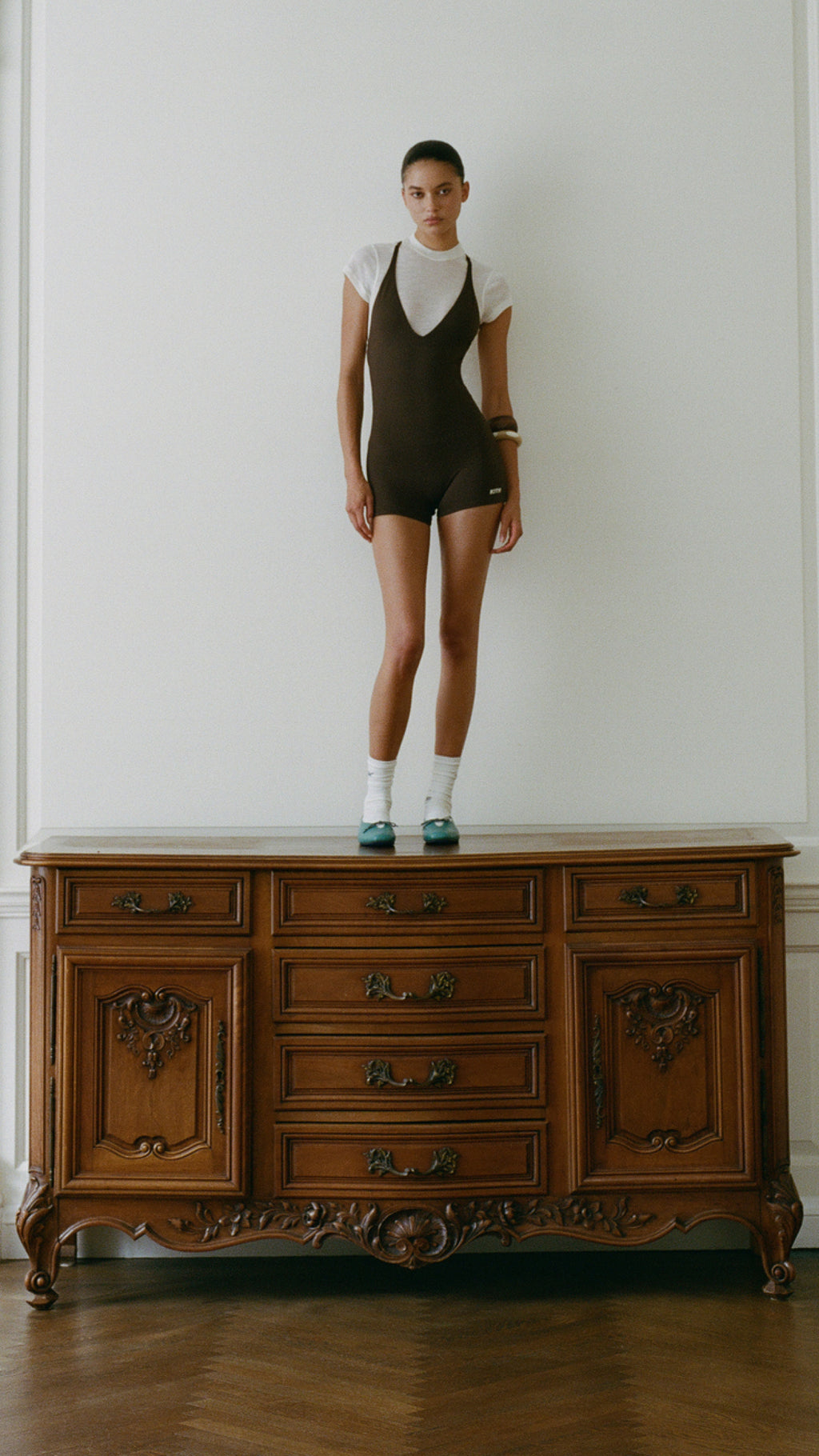 Woman standing atop antique wood dresser wearing white tee under brown Kith bodysuit with wood bead bracelet and teal flats