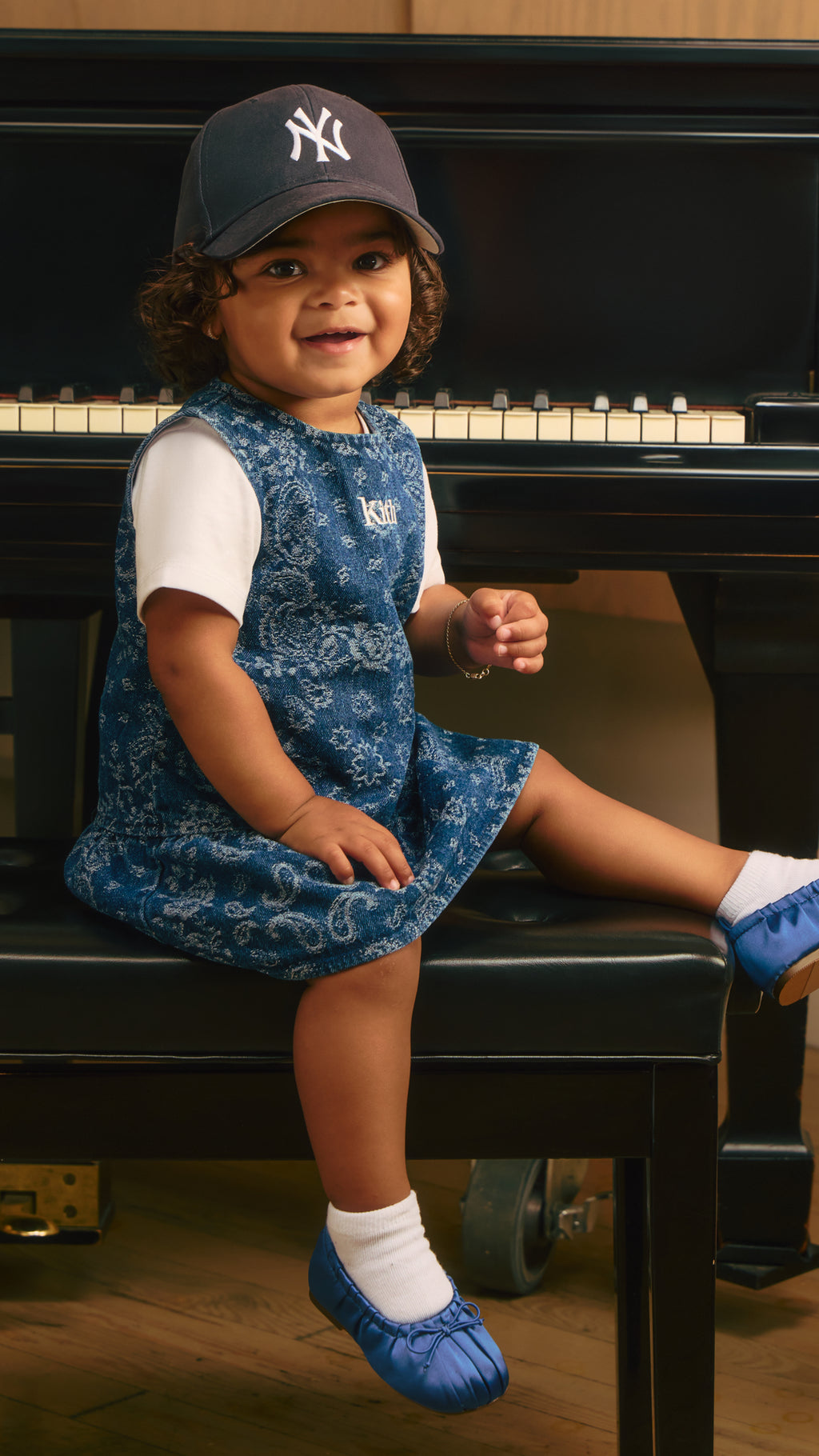 Toddler in a blue floral Kith dress and navy New York cap sitting on a piano bench