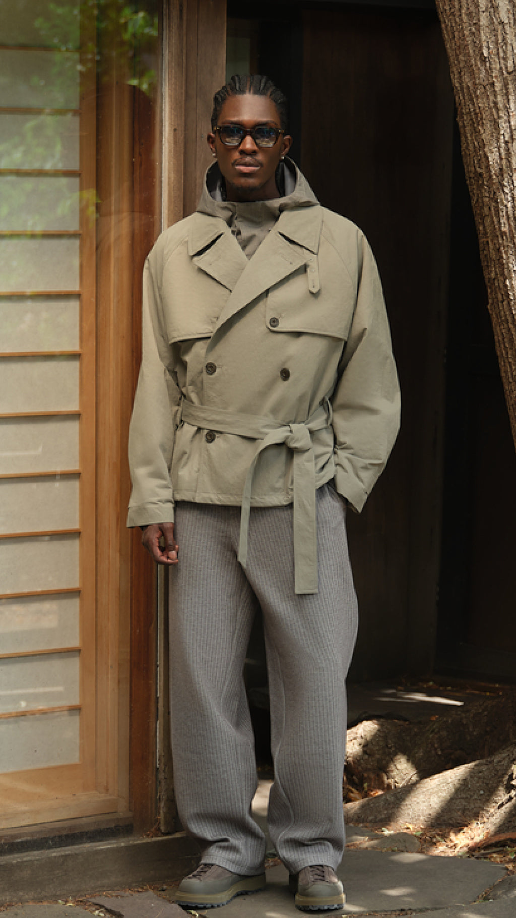Model in an olive double-breasted belted field jacket over a hoodie with grey trousers, vertical crop