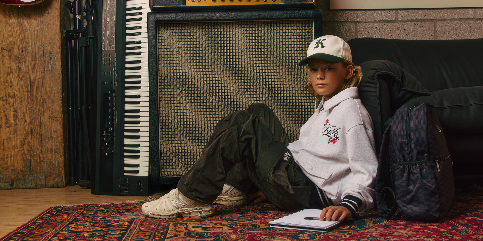 Boy in a black beanie and black Kith crewneck with a white box logo, a backpack strap on his shoulder, gray studio