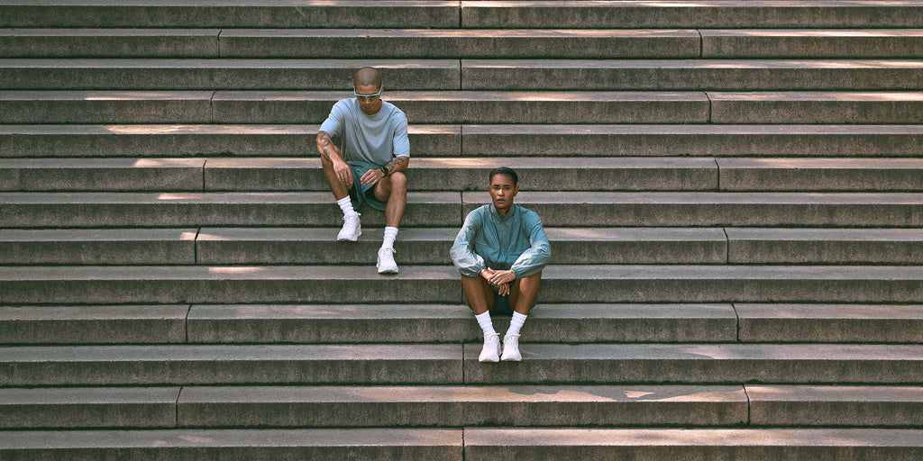Two men sitting on concrete stadium steps wearing pastel blue-green Kith Technology t-shirts, shorts and white sneakers