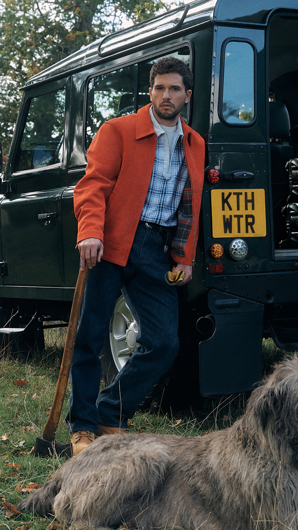 Vertical crop of man in orange jacket and blue plaid shirt against a green Land Rover with plate KTH WTR, holding an axe beside a dog