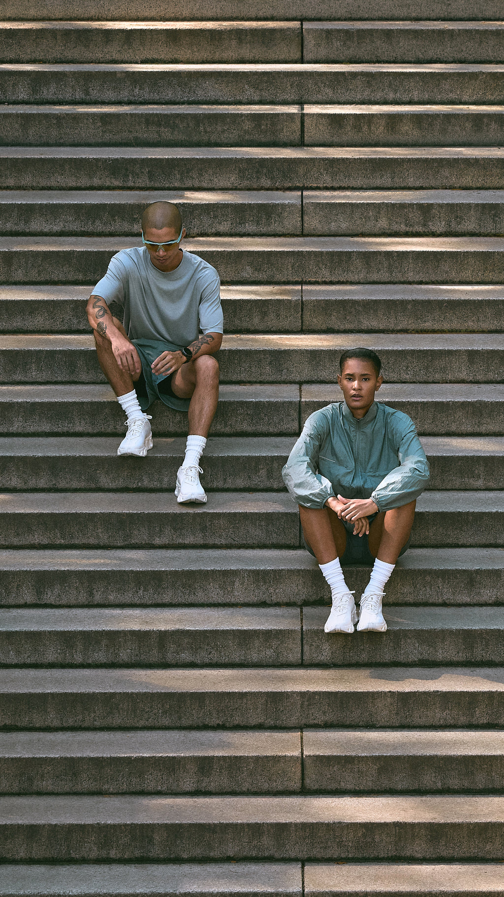 Two men sitting on concrete stadium steps wearing pastel blue-green Kith Technology t-shirts and shorts, mobile crop