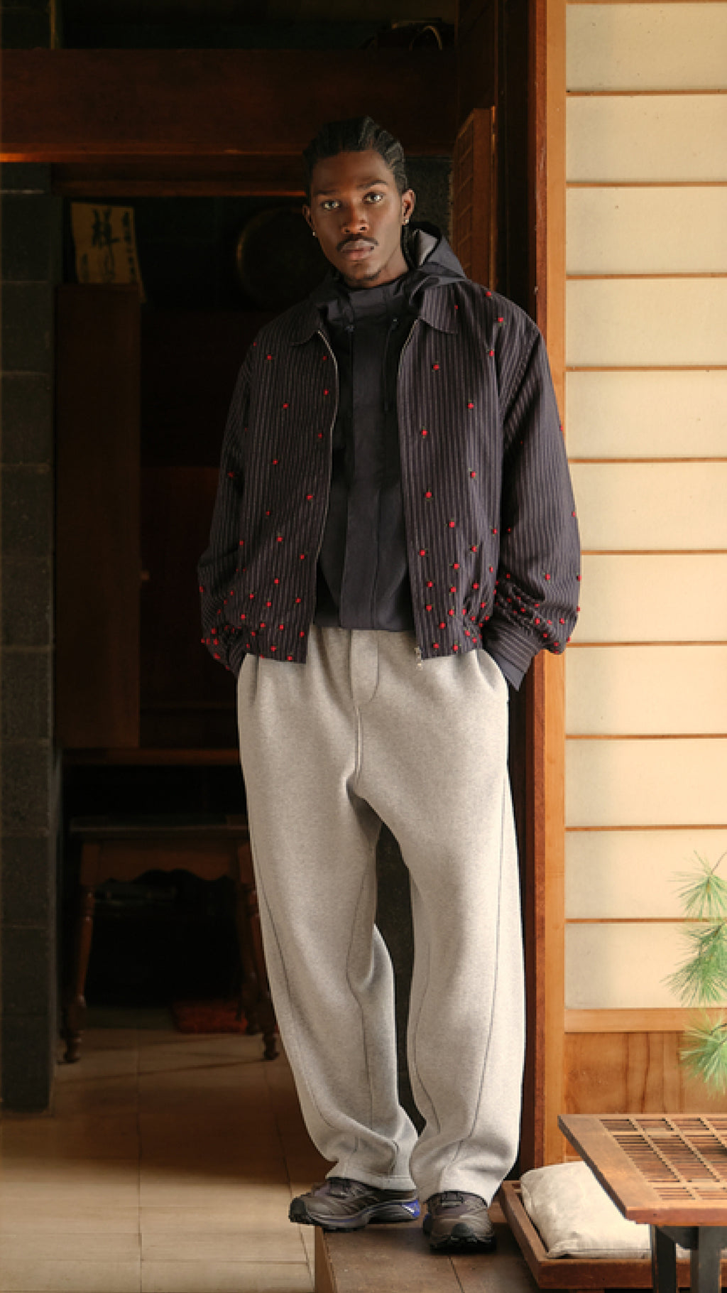 Model in a navy pinstripe bead-embellished jacket over a hoodie and grey sweatpants in a wood doorway, vertical crop