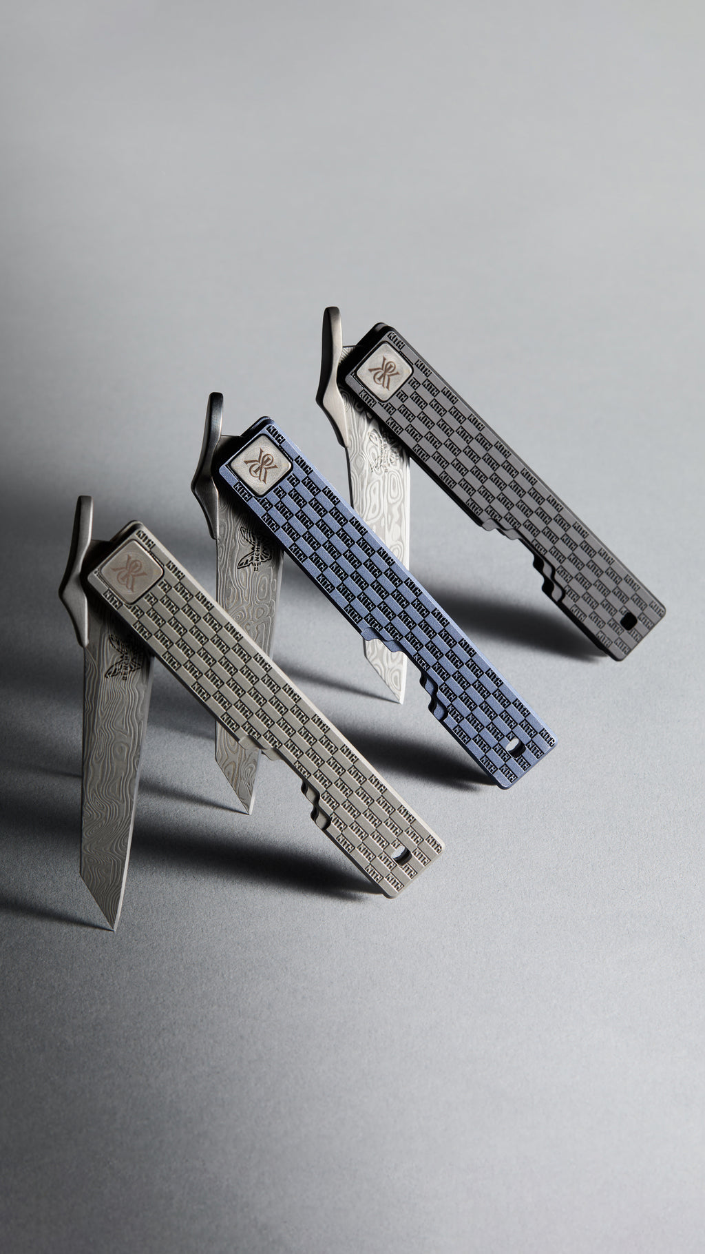 Three Kith x Benchmade folding knives with open Damascus-steel blades and KITH-monogram titanium handles in silver, blue check and grey, vertical crop