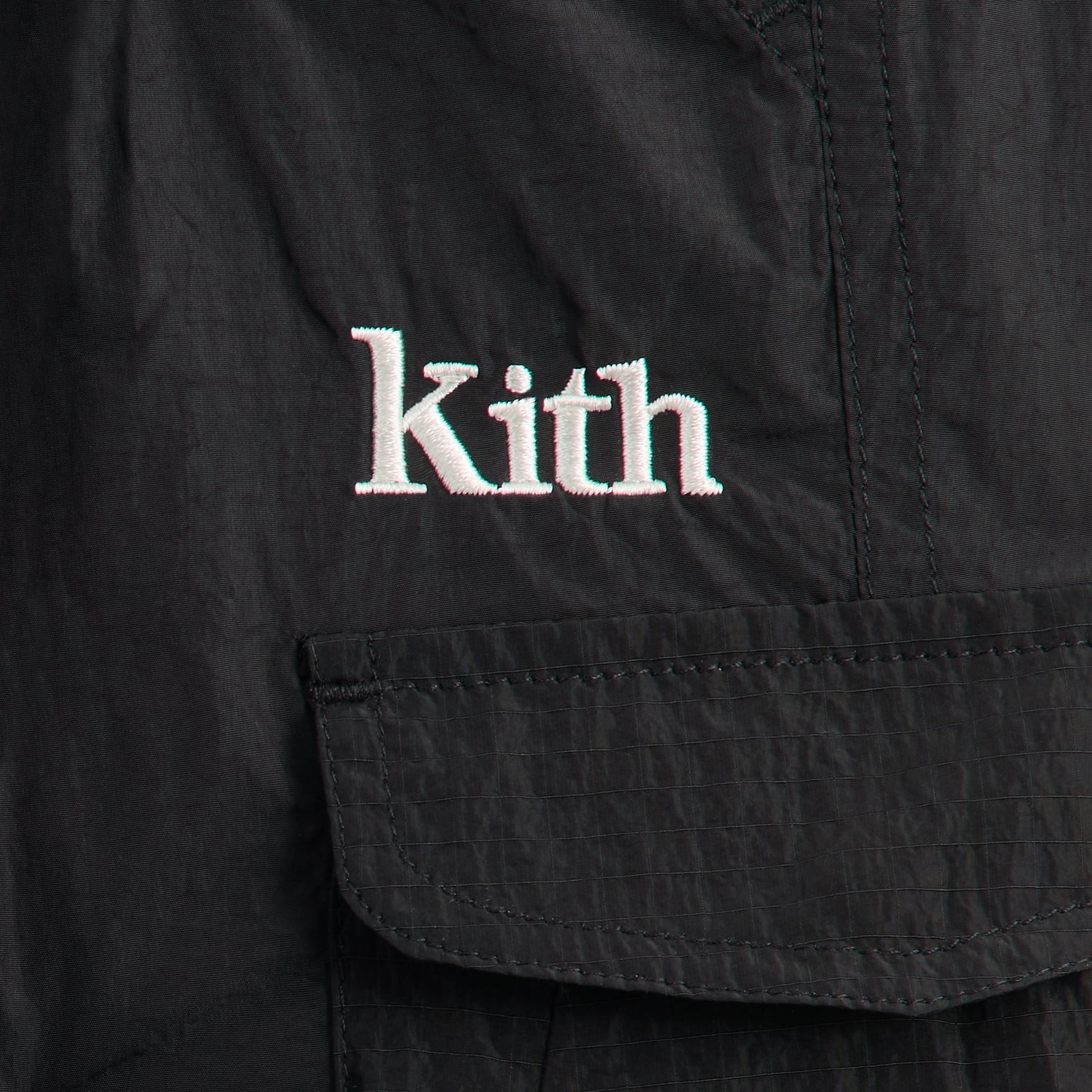 Close-up of white embroidered Kith logo above cargo flap pocket on black nylon parachute pant