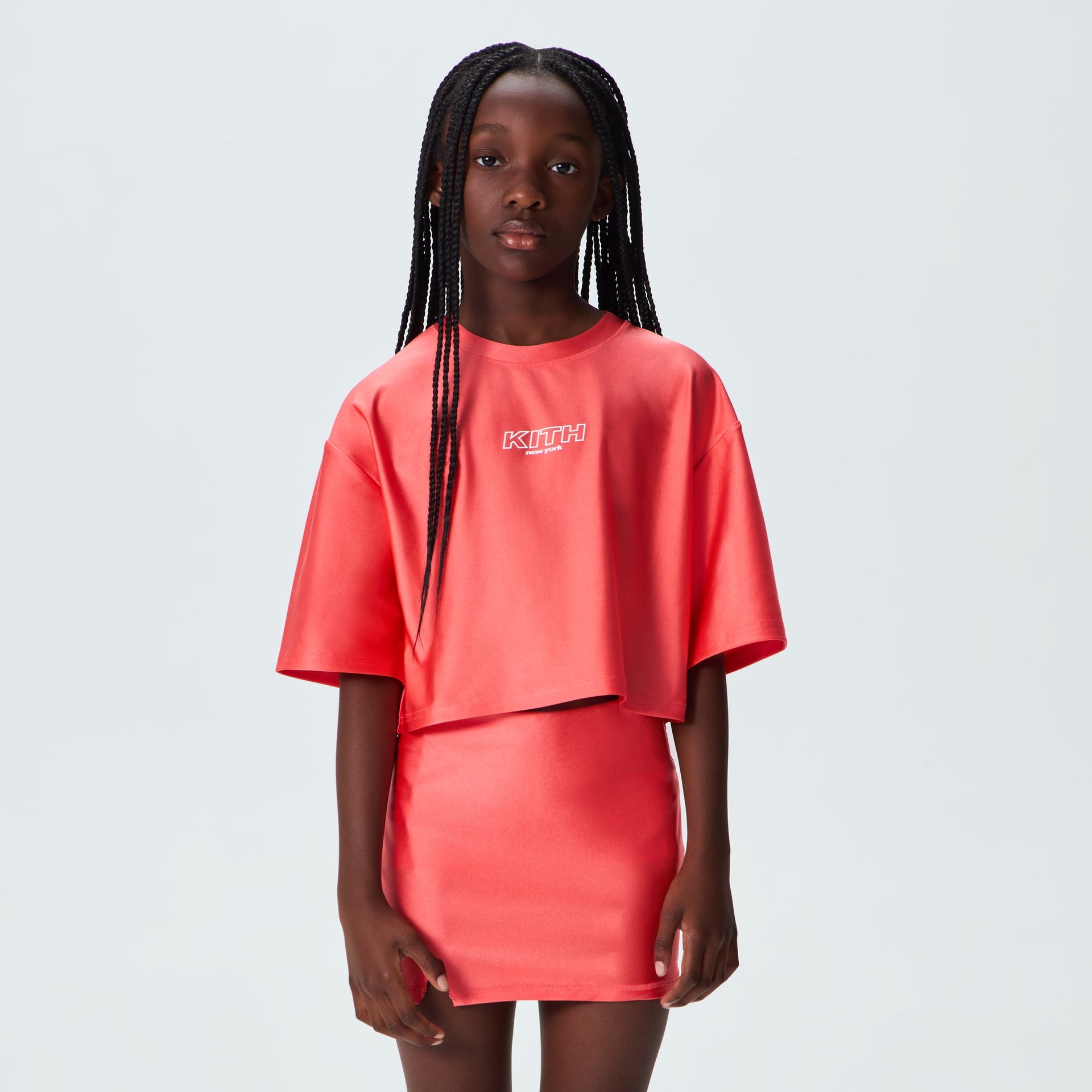 Girl wearing coral Carnation Kith Kids Active Fei Cropped Tee with matching skirt, on-model front view