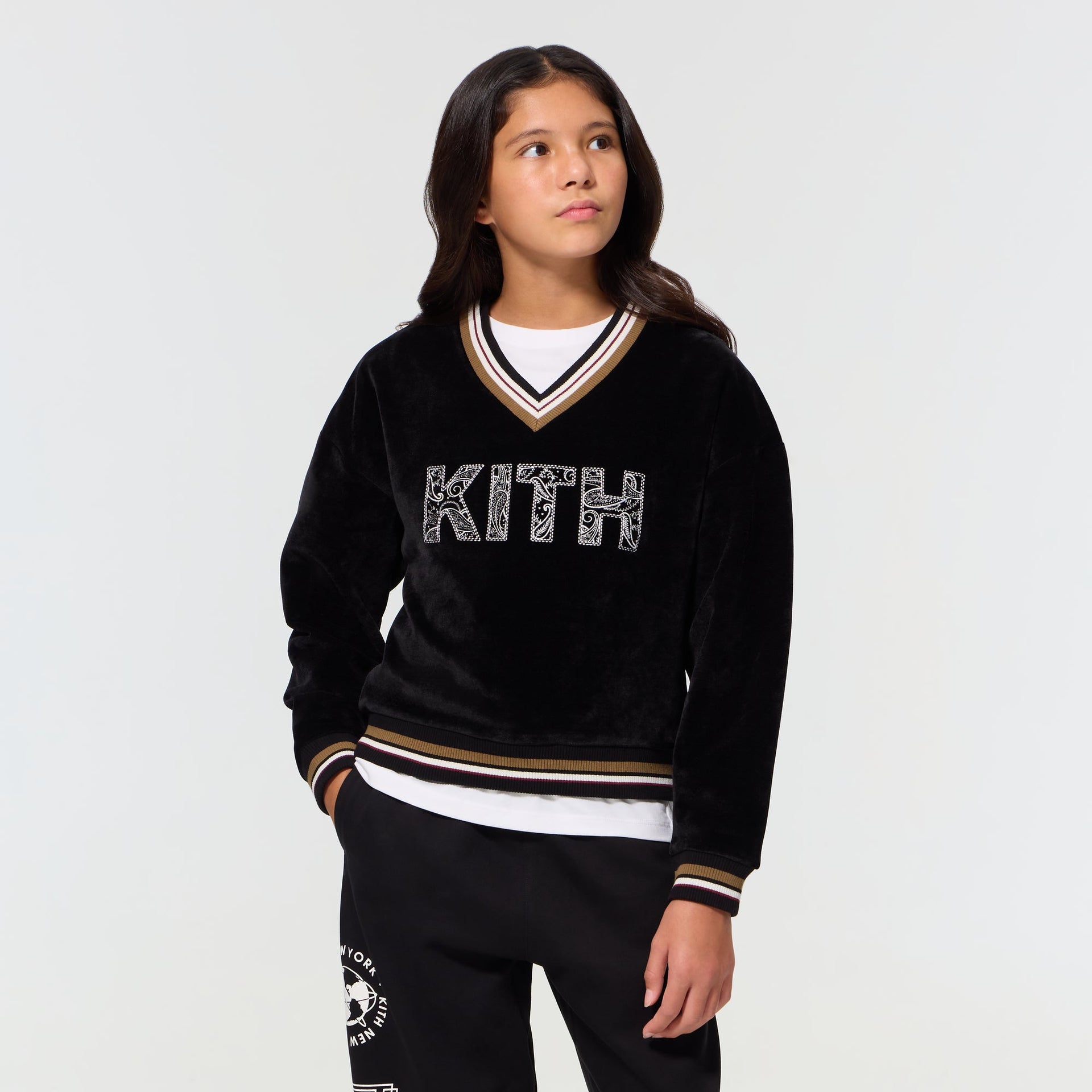 Child wearing the black Kith Kids Textured V-Neck Nelson sweatshirt, front view with striped trim and paisley KITH logo