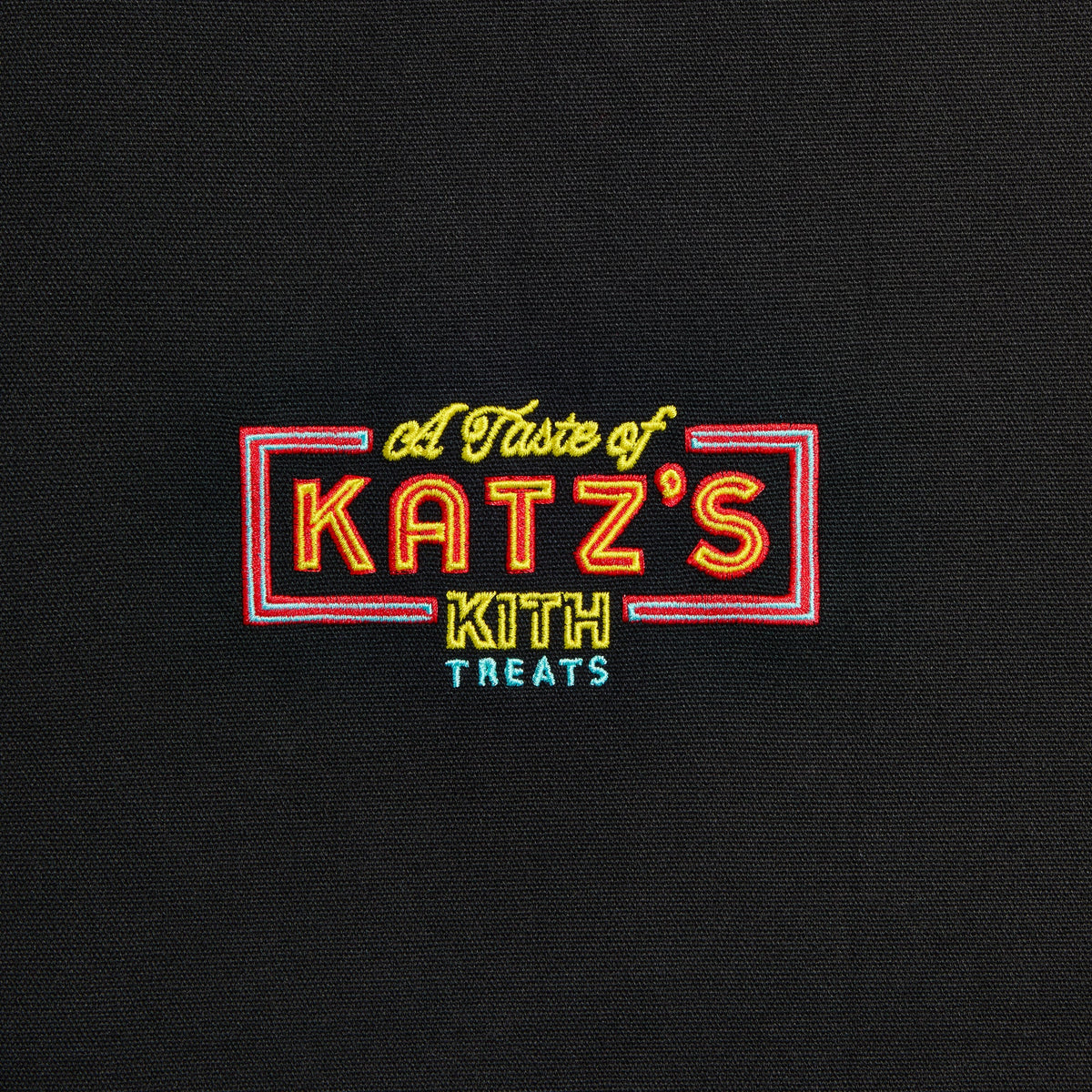 Close-up of red, yellow and blue neon-sign-style 'A Taste of Katz's' Kith Treats logo embroidered on black apron