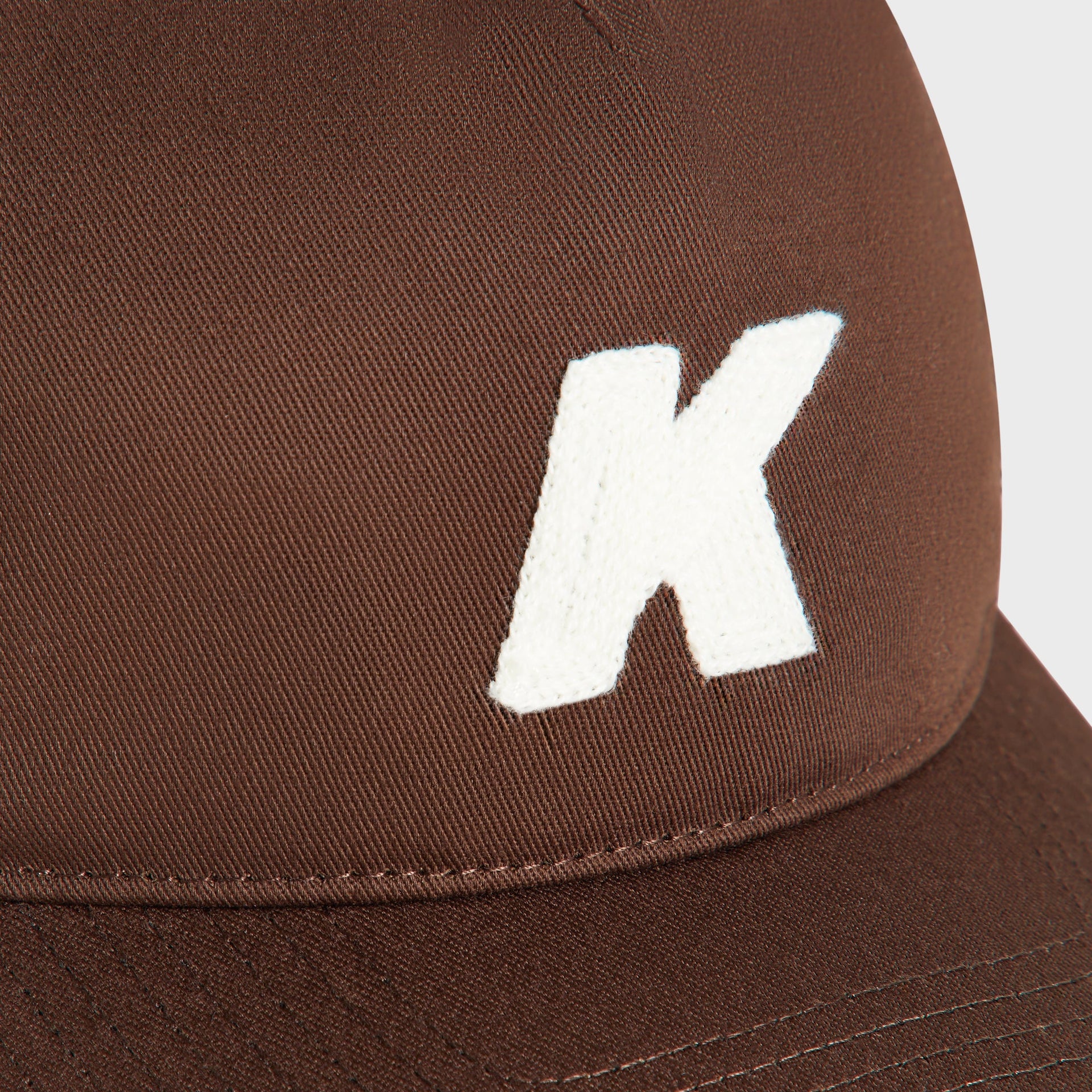 Close-up detail of embroidered white K logo patch on Kith Nolan Trucker Hat in Kindling brown twill