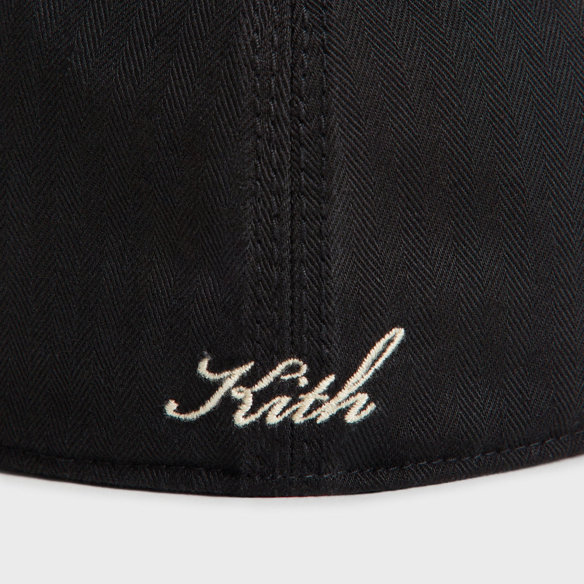 Close-up detail of cream Kith script embroidery near the back seam of the black herringbone Franchise cap