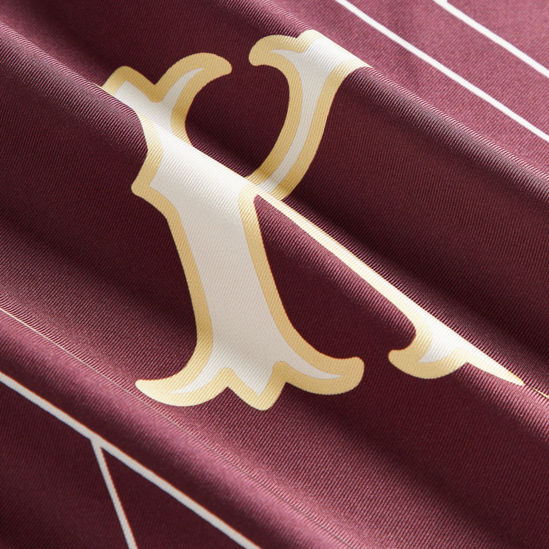 Close-up of the Kith Printed Silk Scarf in Magma showing the gold and cream ampersand monogram on silk