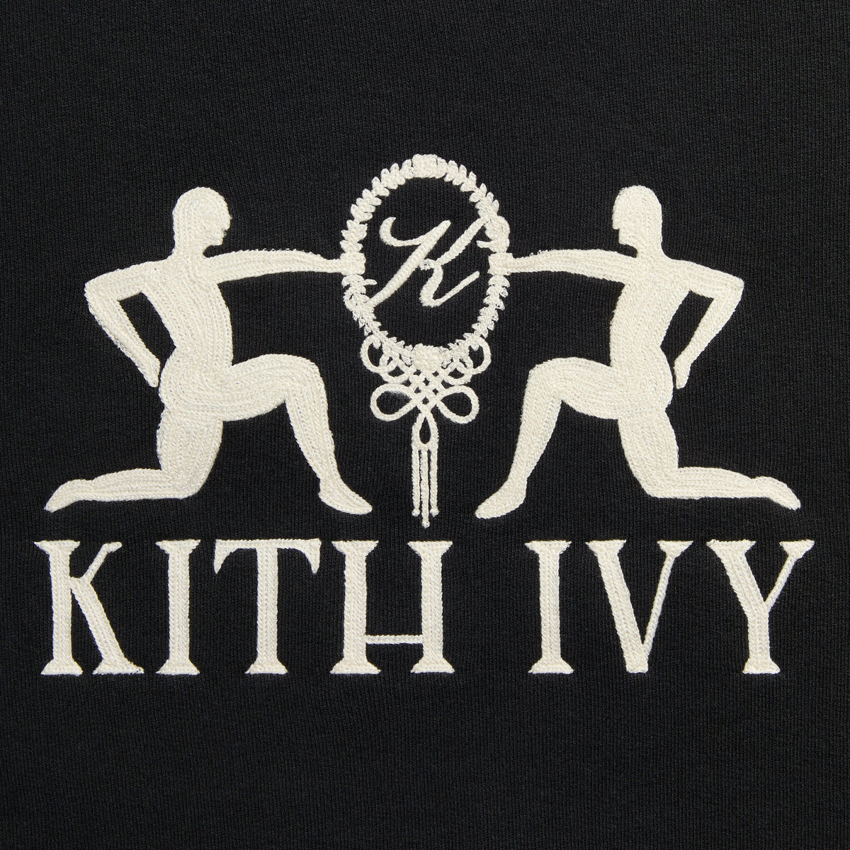 Close-up of cream embroidered Kith Ivy emblem with two kneeling figures and K monogram wreath on black fleece