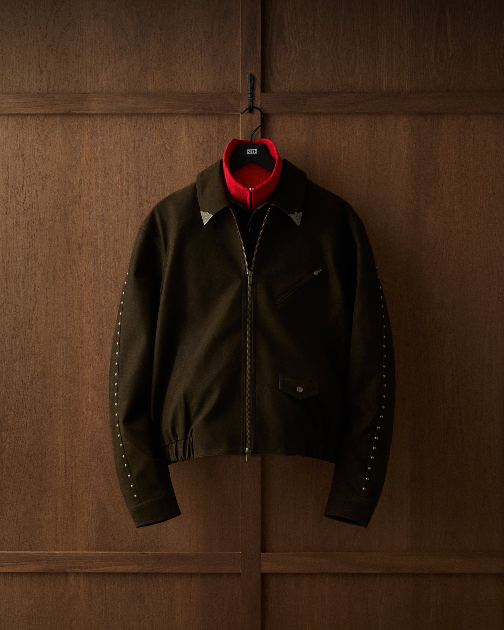 Close-up of a dark olive Kith jacket with silver stud details on the sleeve, hanging against dark wood paneling