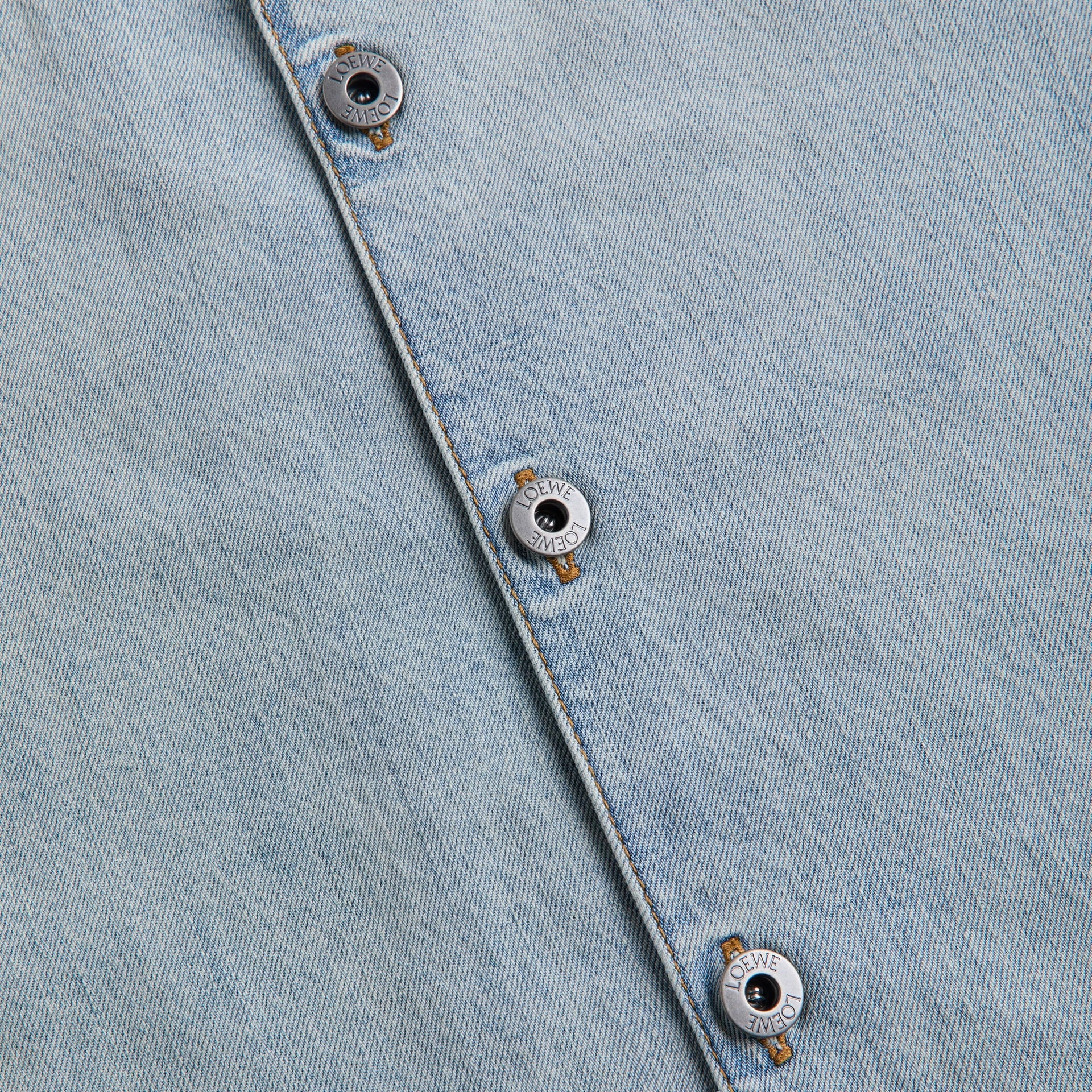 Close-up of engraved silver Loewe buttons on washed light blue denim shirt placket