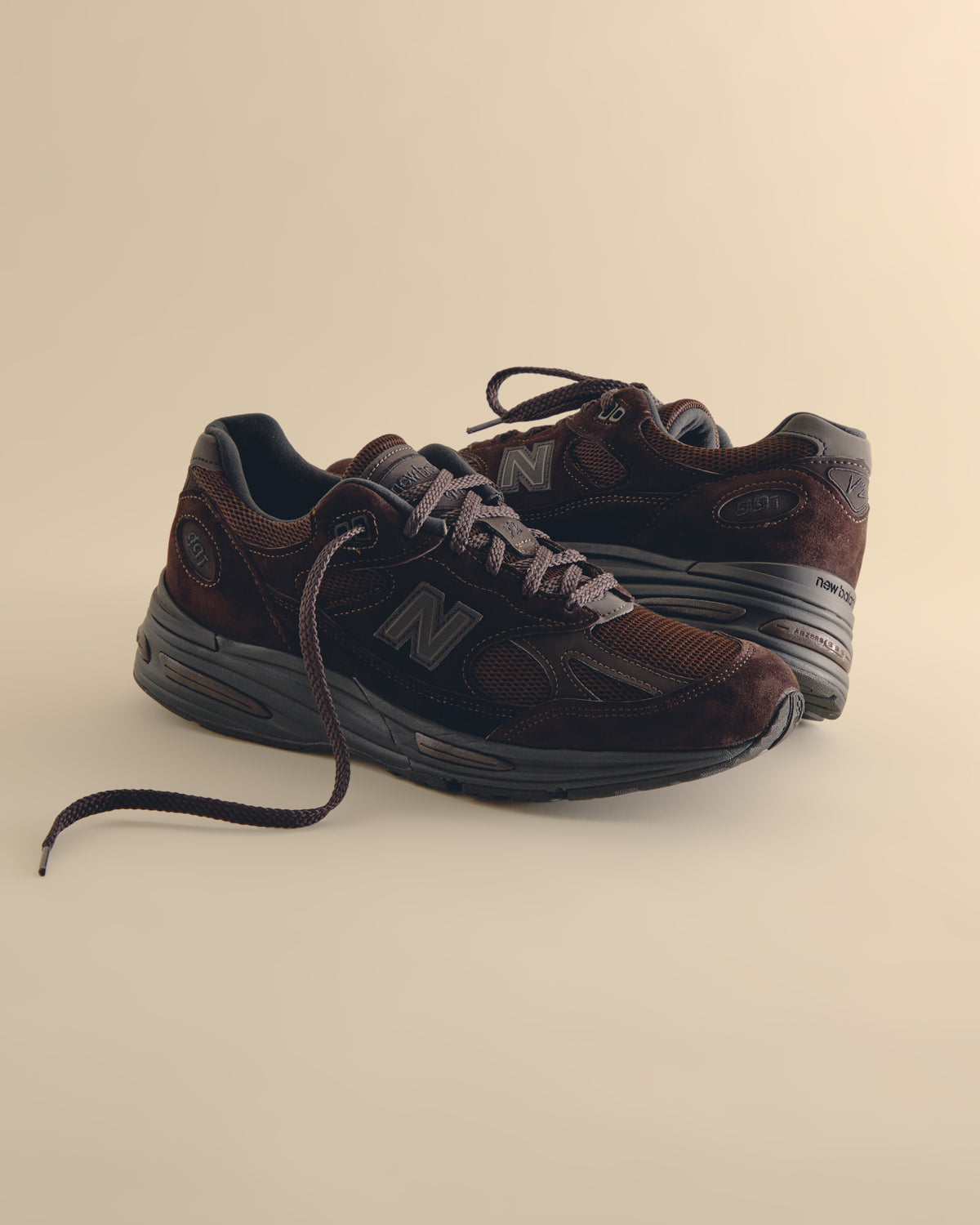 New Balance 991 sneakers in brown suede and mesh with tonal grey N logo and brown laces, pair on tan backdrop