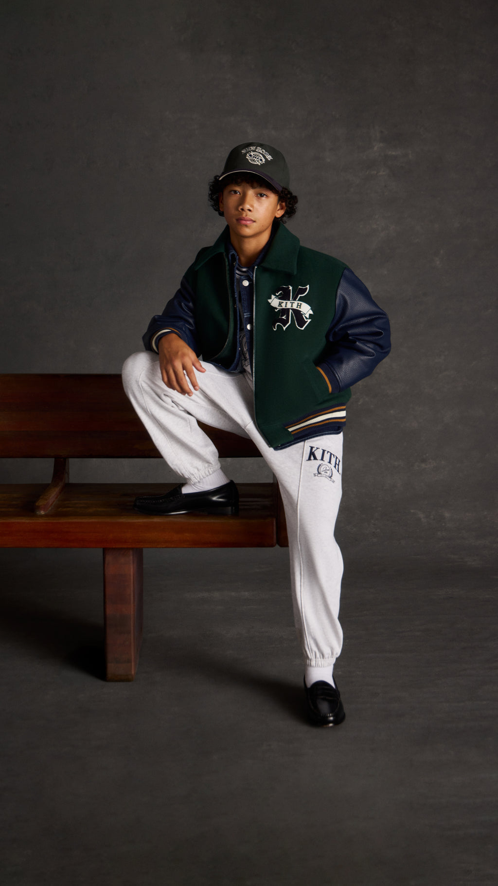 Boy in a green and navy Kith varsity jacket with a K crest patch, cap, and heather grey Kith sweatpants, leaning against a wood bench