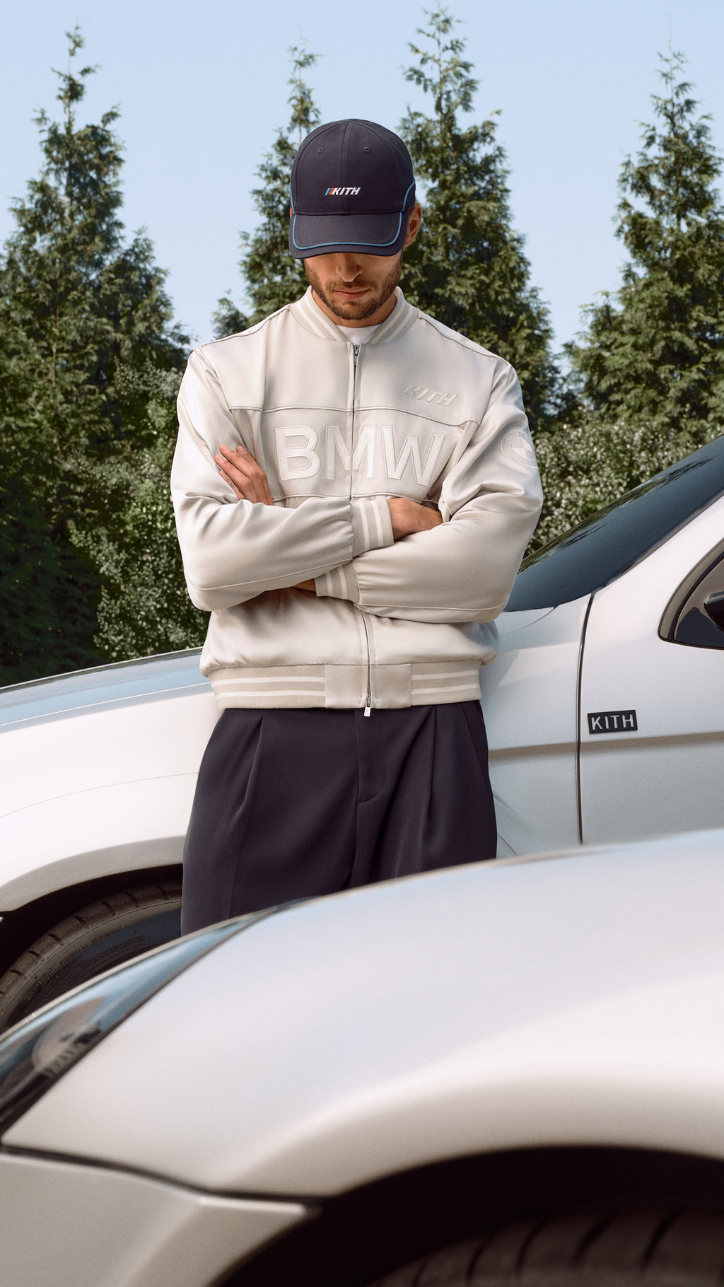Close-up of a man in a white Kith for BMW jacket and navy cap leaning against a white BMW
