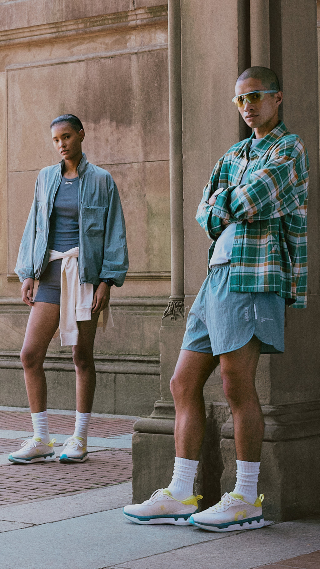 Man and woman at a stone colonnade in Kith activewear, olive jacket with bike shorts and green plaid shacket, mobile crop