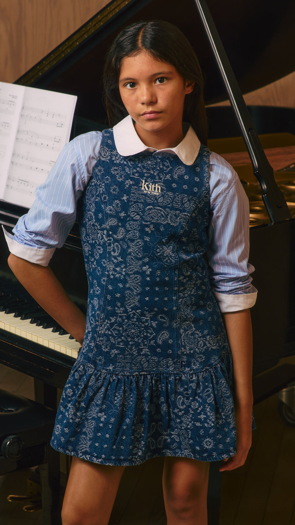 Kith Kids: girl at a grand piano in a blue bandana-paisley Kith New York pinafore over a striped shirt, vertical mobile crop