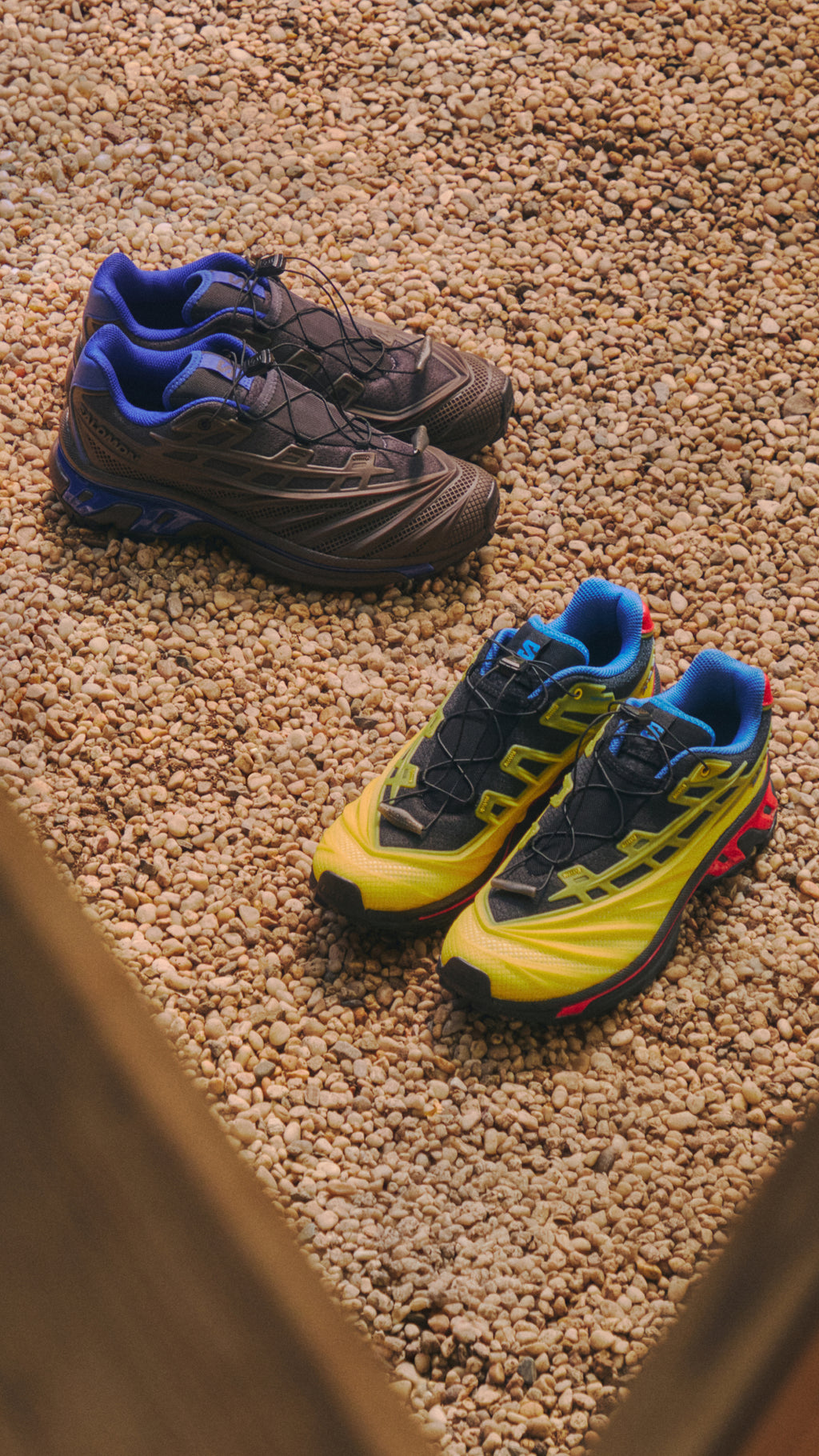 Salomon XT-6 trail sneakers in brown-blue and yellow-black-red set apart on pale pebble gravel, vertical crop