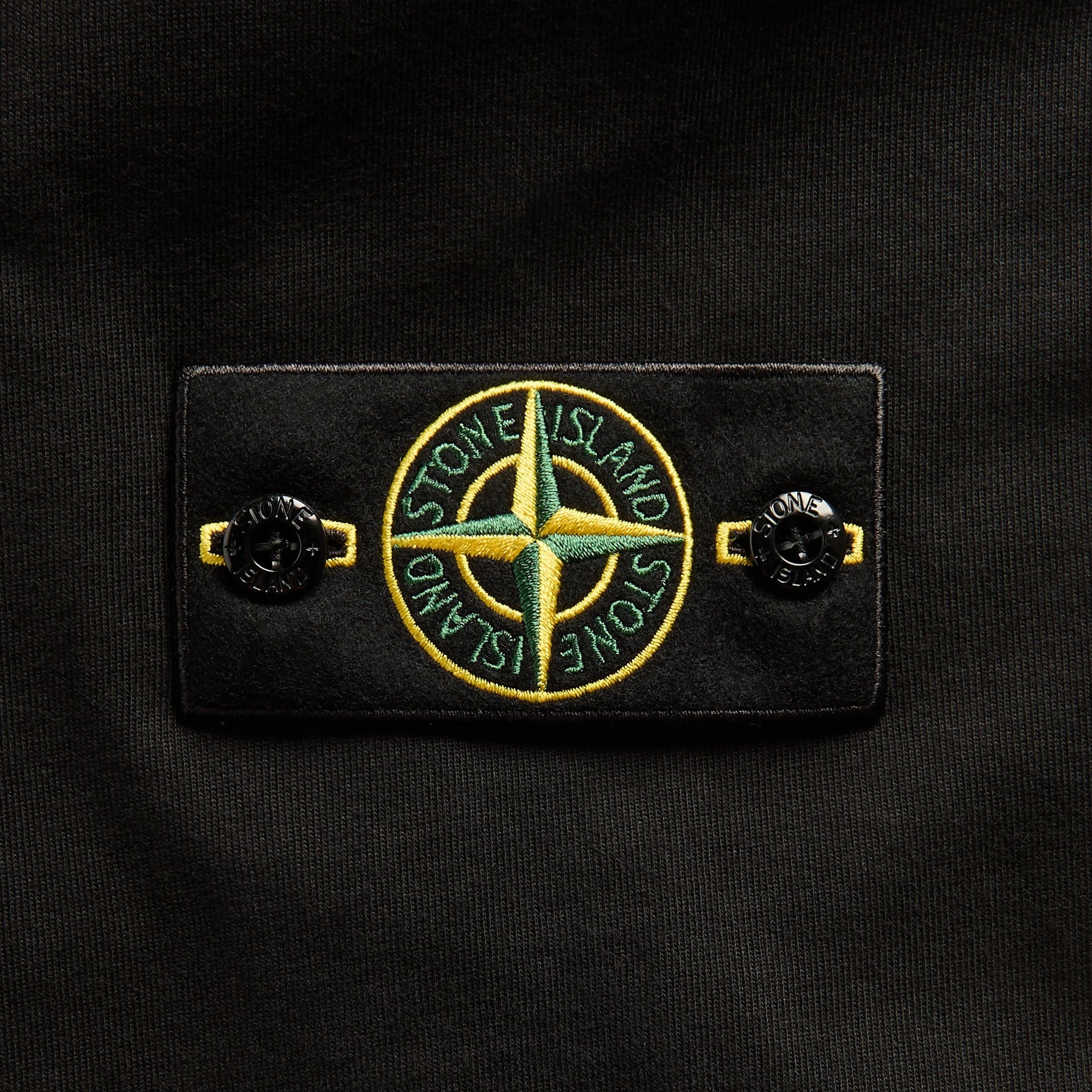Close-up of Stone Island green and yellow compass badge and buttons on black sweatshirt sleeve