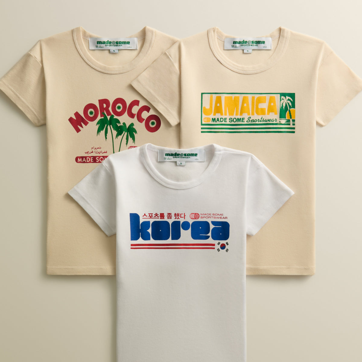 Made Some Sportswear baby tees in cream and white with Morocco, Jamaica and Korea graphics, stacked on a beige backdrop