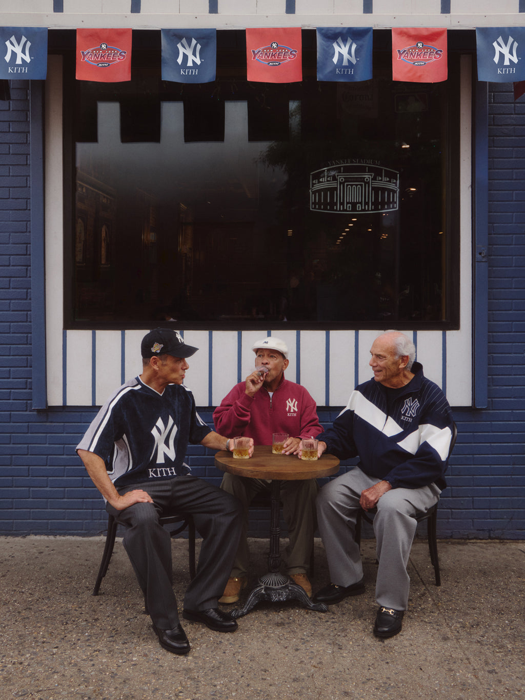Three older men seated at a sidewalk cafe table beneath Kith for New York Yankees banners, wearing Yankees Kith jerseys and jackets