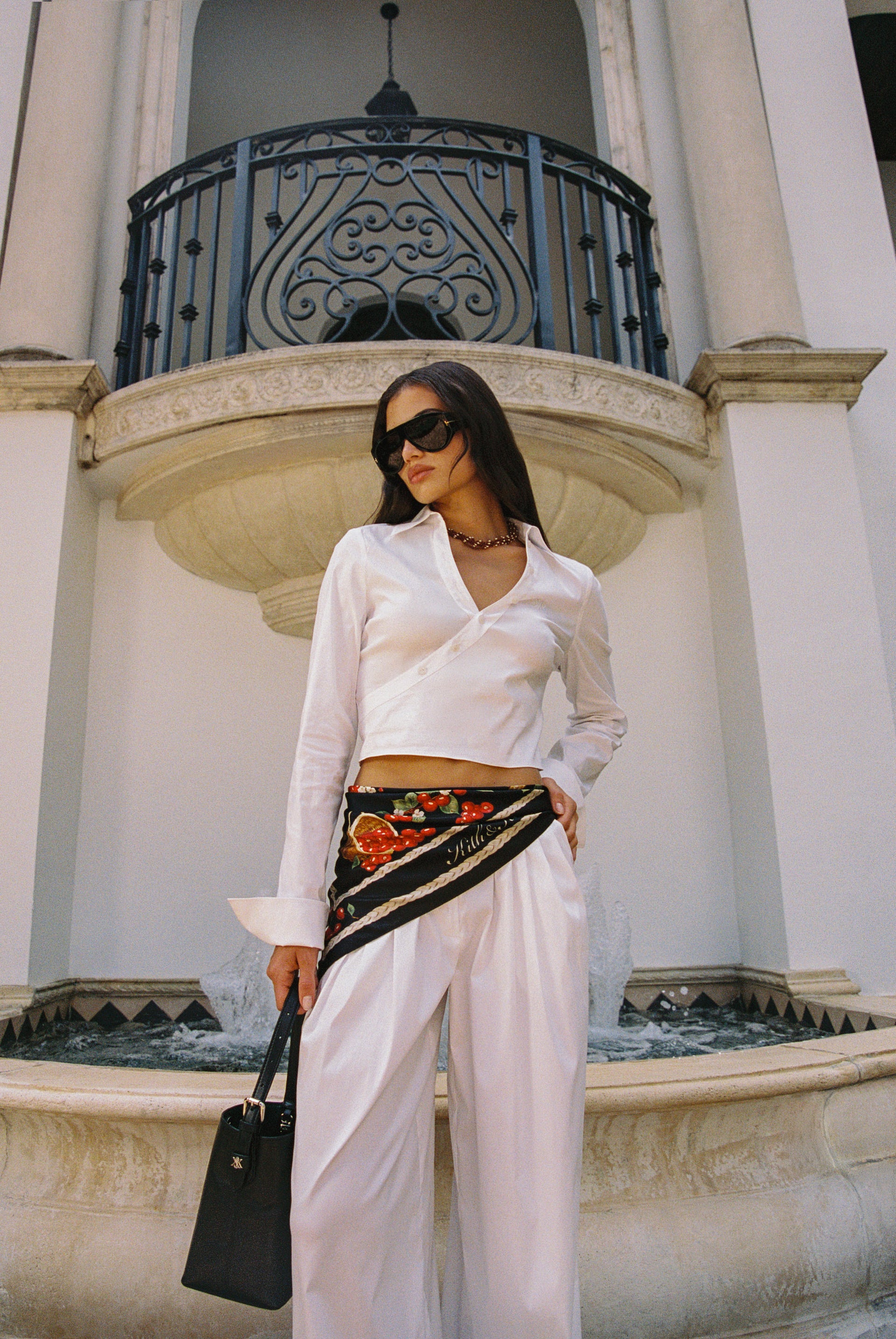 Kith Women Summer 2026: model by a fountain in a white satin wrap crop shirt, wide-leg trousers, baroque waist scarf and black mini bag